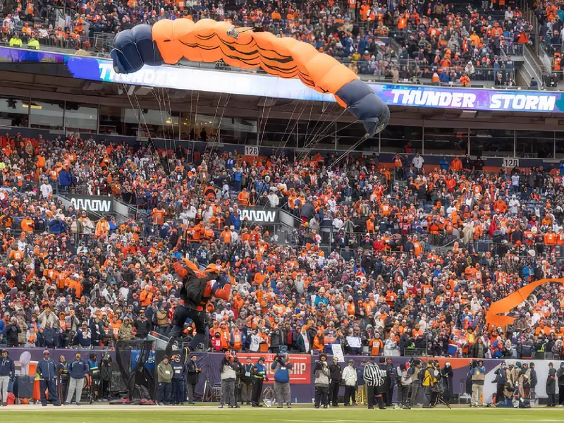 A parachutist landing in front of a packed orange-clad crowd in a football stadium