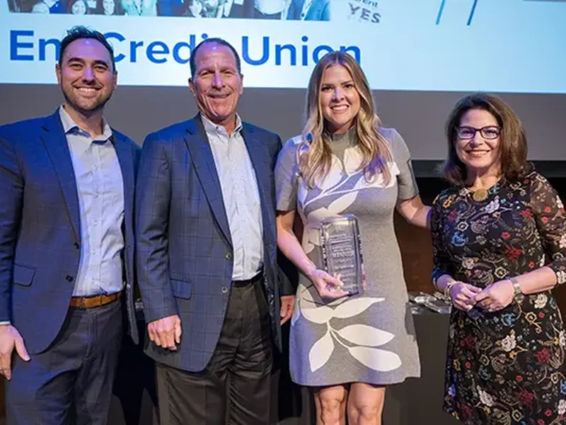 Four people posing on a stage with a woman holding a glass award in front of an Ent Credit Union screen