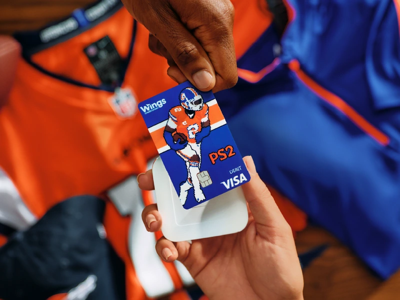 A hand holding a Wings Credit Union debit card with a pixel-art football player over orange and blue jerseys