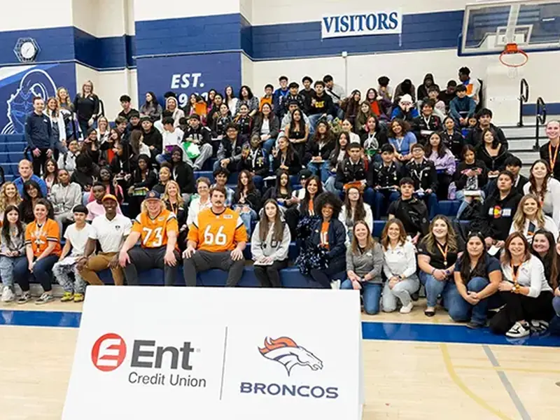 A large group of students posing in a gym with two football players behind an Ent Credit Union and Broncos sign