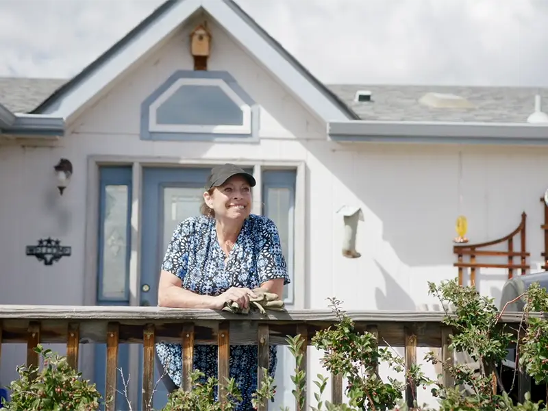 Woman leaning on wooden porch railing in front of light-colored house with plants and trellis