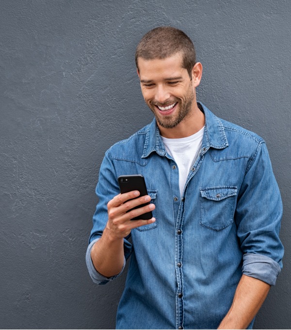 Cool, casual and smiling man looking down at his smartphone as he leans back against a wall. He's excited to find out about Ent Extras Checking.