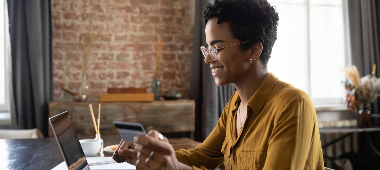 Happy young African woman using laptop and credit card to review finances online.