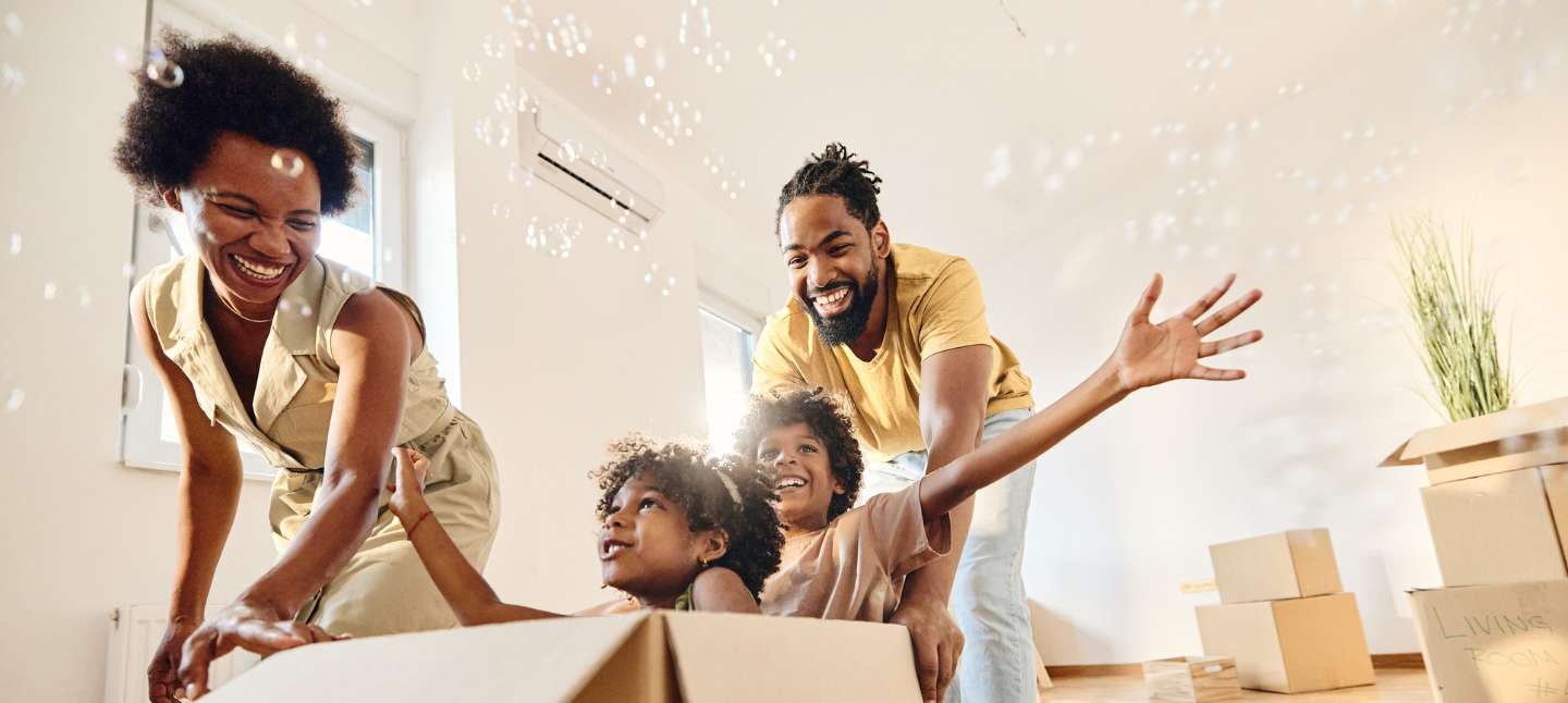Playful African American parents having fun while pushing their small kids in carboard box at new house.