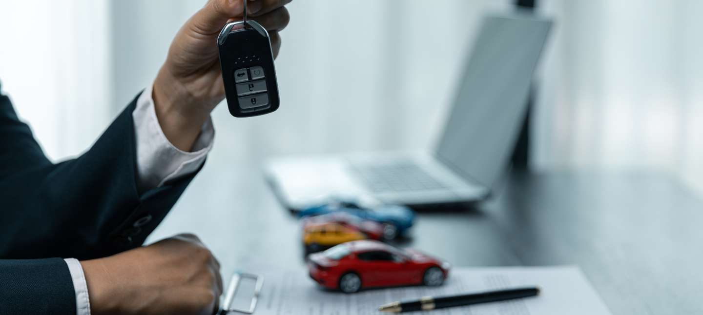 Closeup of car sales person's hand holding a car key with the car contract and paperwork laid out on the desk in front of them.