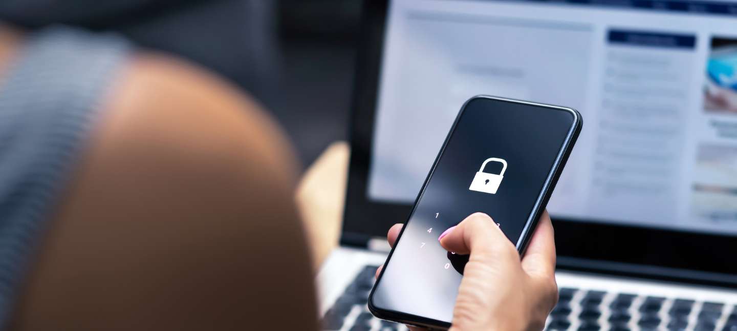 Over-the-should view of an individual holding a smartphone with password screen lit up. Laptop open in the background.