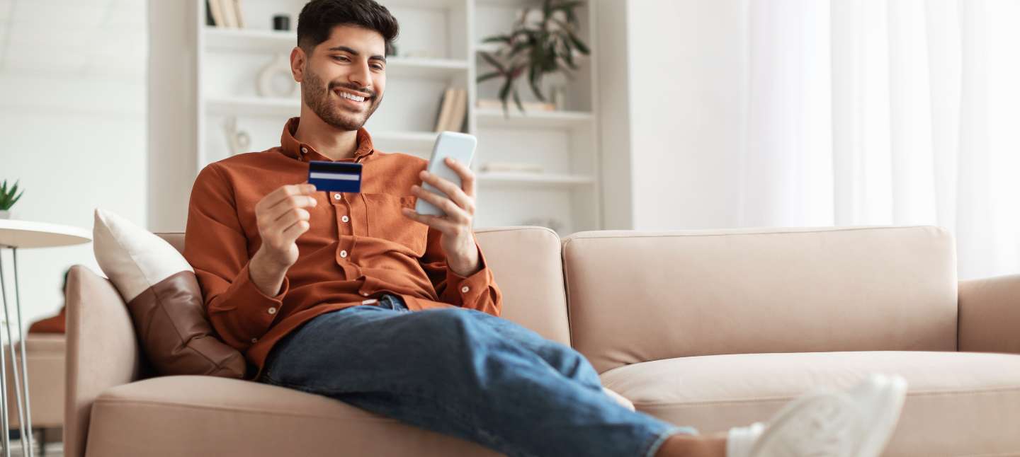Smiling young man holding debit credit card in hand and using cell phone, making financial transaction while sitting on the couch at home in living room.