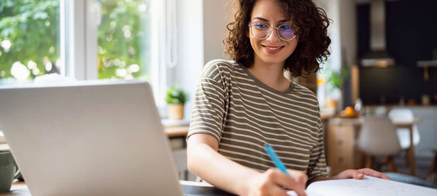 Young woman smiling at laptop while writing in her notebook.
