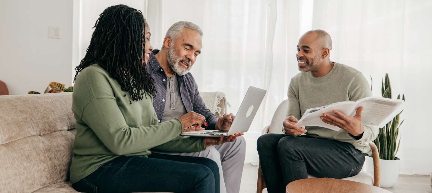 A couple on a couch looking at a laptop while consulting with a man with a folder of paperwork