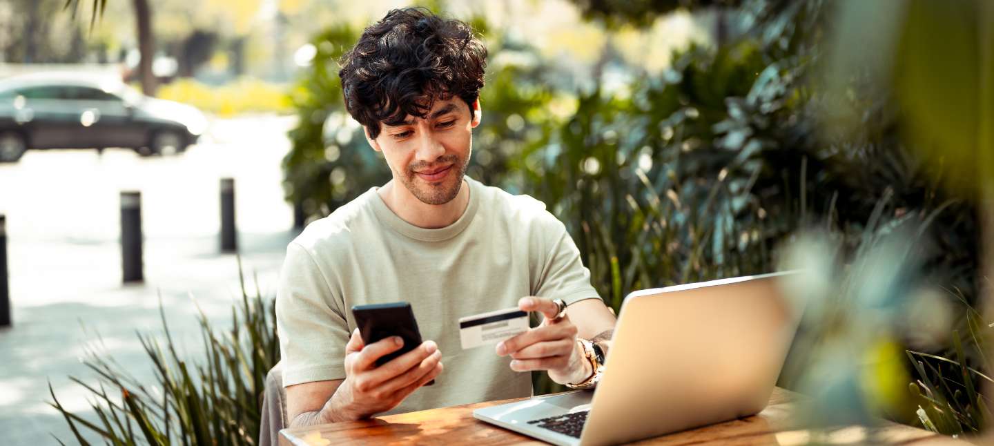 Caucasian young man sitting in front of a computer while holding his phone and a debit card