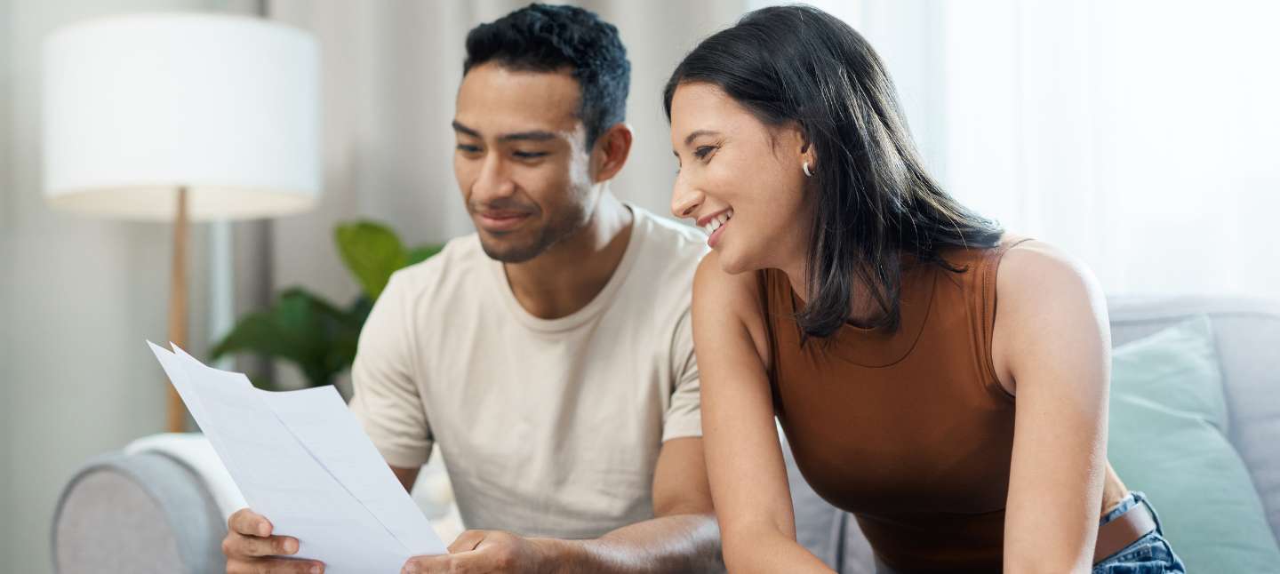 Man and woman smile while sitting on the couch and holding financial paperwork.