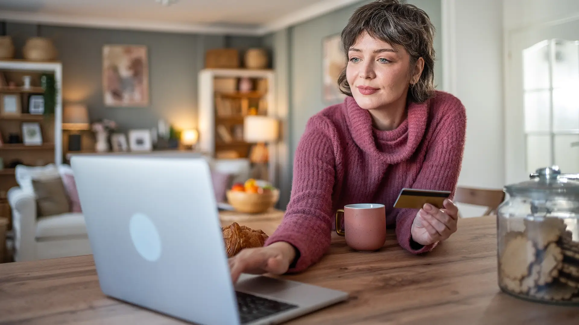 A woman sits at the kitchen counter and checks her credit card, using a digital tablet