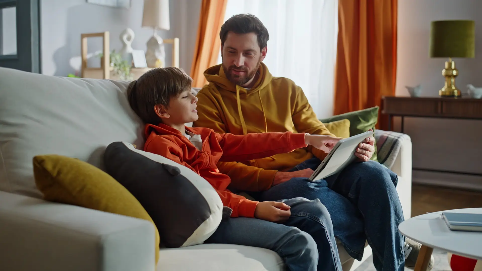 A father and son sitting on the couch talking and using a tablet